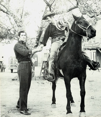 Dressed in his civvies, Clint (“Cheyenne”) Walker shakes hands with Ty Hardin in costume for his “Bronco” series on the Warner Bros. back lot in 1958. (Thanx to Neil Summers.)
