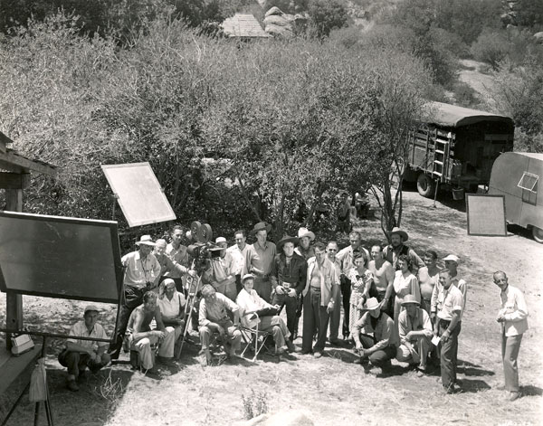 Cast and crew of “Frontier Revenge” (‘48) assemble on Iverson’s Ranch for a group photo. Lash LaRue dead center, to his right is Jim Bannon. Is that producer Ron Ormond to Lash’s left, shirt open? Director Ray Taylor and most likely script supervisor Moree Herring are seated to Lash’s right. Co-stars Ray Bennett and Lee Morgan are in the back to Lash’s left. Others unidentified. Can anyone help? Cute to notice the kids looking on while hiding in the bushes. Obviously missing from the cast foto are Fuzzy St. John, Peggy Stewart and Sarah Padden.