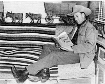 Touring the country with his Bar Bar A Ranch Rodeo, Bill Elliott relaxes in his deluxe house trailer. Covered wagon scale model wood carving in the background was given to Bill by famed sculptor A. Lee Norvell. 