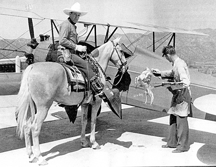 At Burbank, CA’ airport in 1931 Ken Maynard and Tarzan admire the latest edition to his fleet of seven airplanes, this one a Stearman bi-plane. The artist is Harold E. Bishop. The caption reads, “Let ‘er Buck”. 