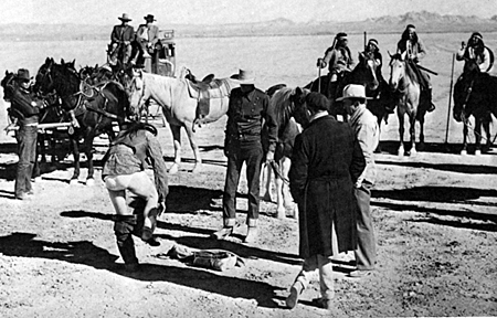 John Wayne watches as Yakima Canutt gets into his Indian costume for the famous leap from a running horse to the lead horse of the stagecoach team at 45 miles an hour. Man in the overcoat is “Stagecoach” director John Ford. 