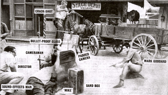 How it’s done. Mark Goddard, Culley on “Johnny Ringo”, blazes away while the cameraman, the director and a sound effects man cower behind sand bags and bullet proof glass. Extra bullet holes are provided by crack shot Jim Rug.