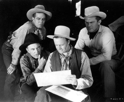 Hoot Gibson, Harry Carey and Guinn “Big Boy” Williams watch Wally Wales put finishing touches on a western landscape painting during a break in the filming of “Powdersmoke Range” (‘35).