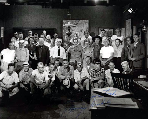 For Jimmy Wakely’s last Monogram western in 1949, “Lawless Code”, this partial cast and crew photo was taken. You can easily spot Jimmy, director Oliver Drake (with mustache and open shirt collar behind Jimmy), Myron Healey to the right of Ollie, leading lady Ellen Hall to the left of Ollie, Kenne Duncan to Ellen’s left, Steve Clark to Ollie’s right in dark coat, Terry Frost and Tris Coffin standing to the right. Set continuity lady Helen McCaffrey is to the right of Steve Clark. We’re also sure Assistant Director Eddie Davis and Cinematographer Harry Neumann are in the group but unsure which two they may be. A good guess would be the man kneeling to the left of Jimmy and the man standing to the right of Drake. Note Wakely’s autograph on the photo. (Photo courtesy Bobby Copeland.)