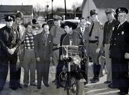 Fuzzy St. John seems to be saying, “How come you get to ride on the motorcycle?” Unsure of the city in which this law enforcement group photo was taken. (Thanx to Jerry Whittington.)